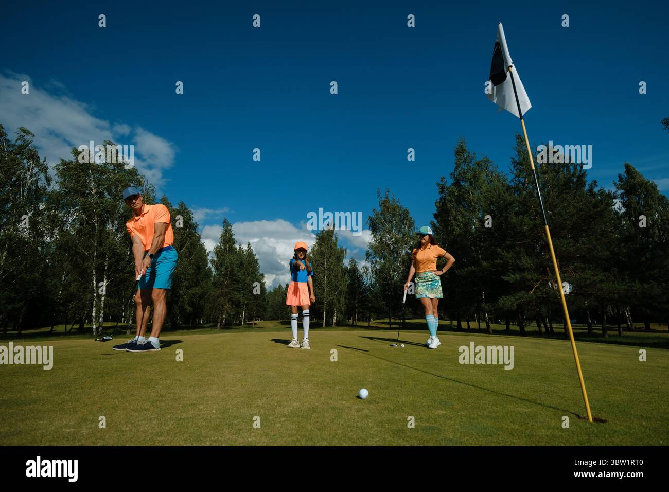 Drei Familienmitglieder nehmen an einer lustigen Golfsession auf einem pulsierenden Golfplatz unter klarem blauen Himmel Teil. Eine Person setzt, während andere beobachten und anfeuern, Stockfoto