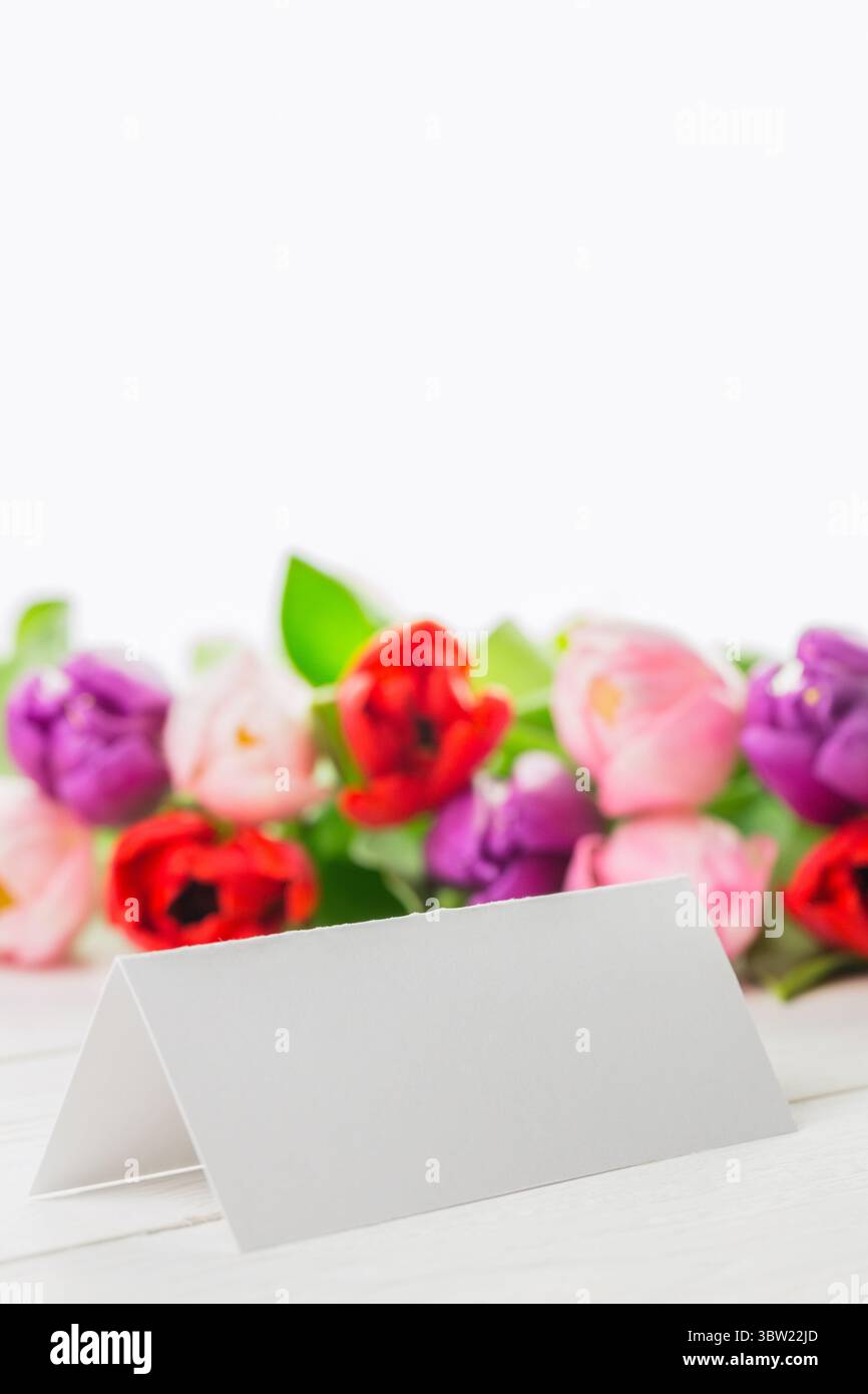 Leere weiße Zeltkarte liegt auf hölzernen Tischplatten in flachem Mockup, mit verschwommenem Tulpenhintergrund Stockfoto