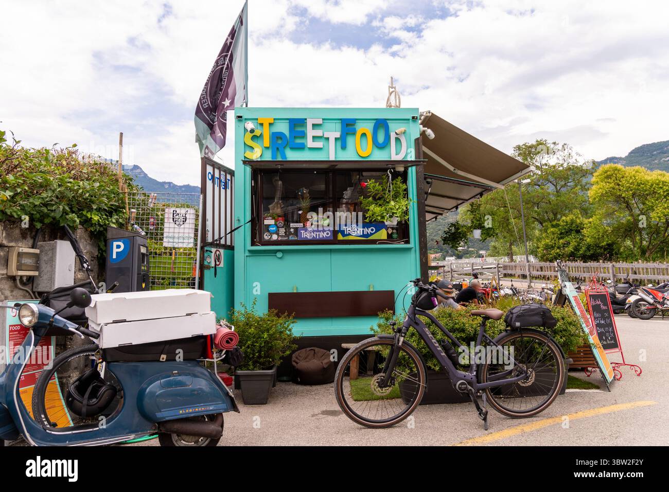 Nago-Torbole, Trentino, Italien - 15. Juli 2025: Gipsyvan in Torbole am Gardasee serviert STREETFOOD in grünem und blauem Wohnwagen mit großem STREETFOOD-Schriftzug. Davor befinden sich ein E-Bike, ein Pizzaroller und Sitzplätze für Gäste *** der grün-blaue Anhänger mit großer STREETFOOD-Beschriftung, Gipsyvan in Torbole am Gardasee in Italien serviert Streetfood. Davor stehen ein E-Bike, ein Pizza-Motorroller und Sitzmöglichkeiten für Gäste Stockfoto