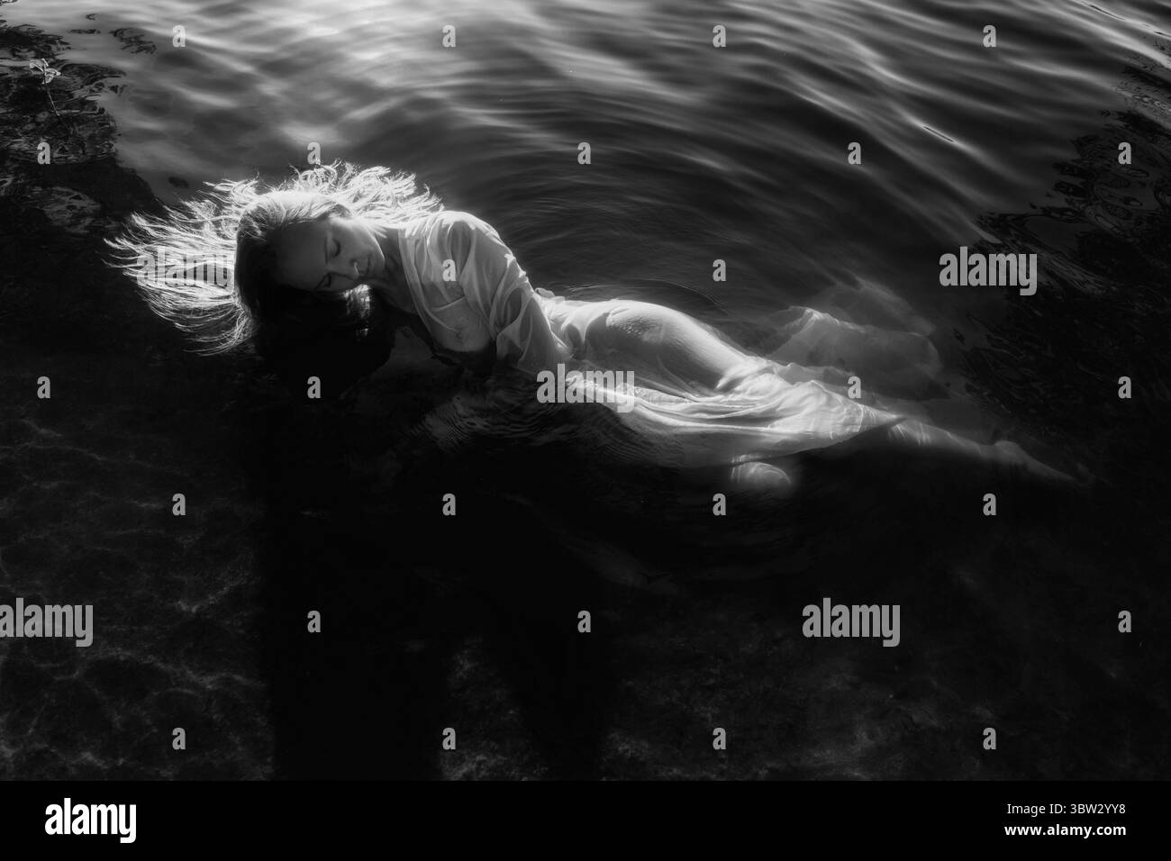 Eine Frau mit langen Haaren liegt sanft auf der Wasseroberfläche und trägt ein fließendes Kleid. Sanfte Wellen umgeben sie, während sie einen friedlichen Moment genießt Stockfoto