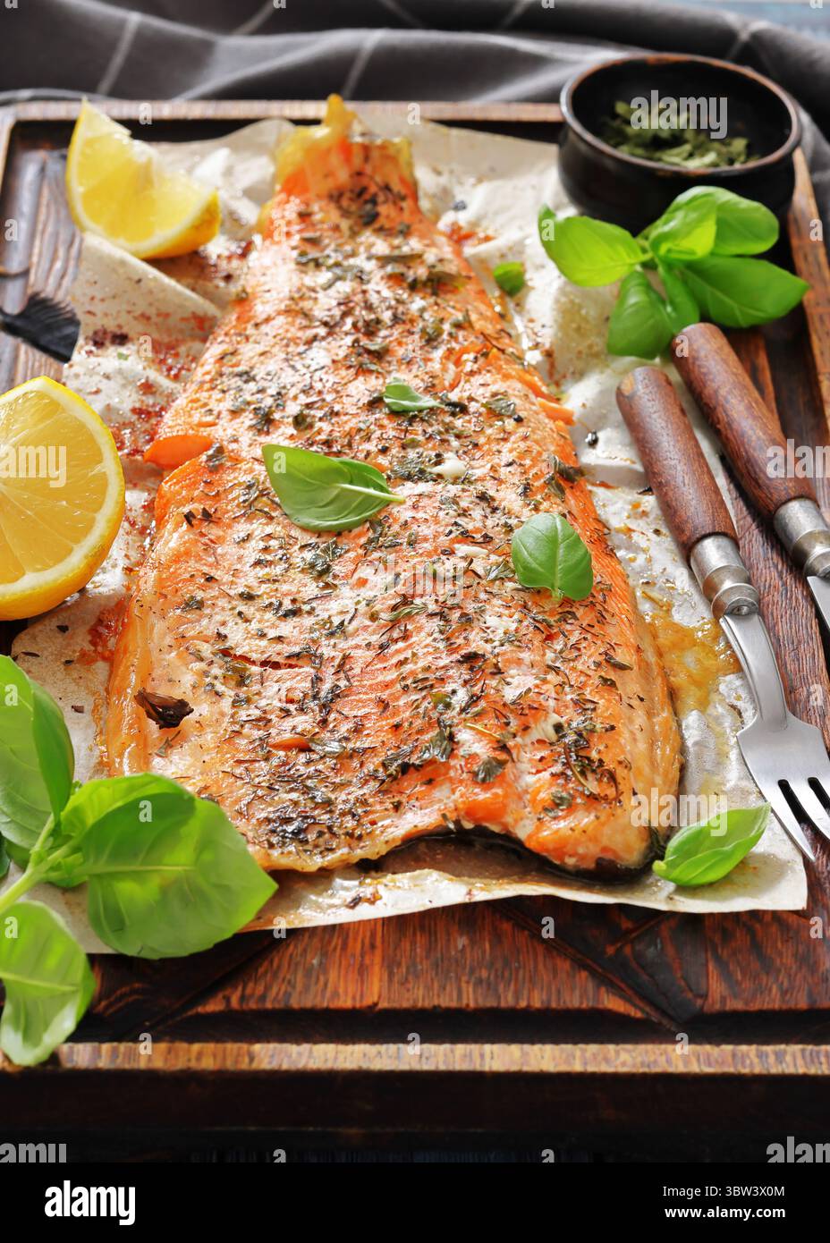Gegrillte ganze Lachsfilets mit Haut auf einem rustikalen Holzschneidebrett mit frischem Basilikum, Zitronenscheiben, Besteck und Gewürzen auf Holztisch, vertica Stockfoto