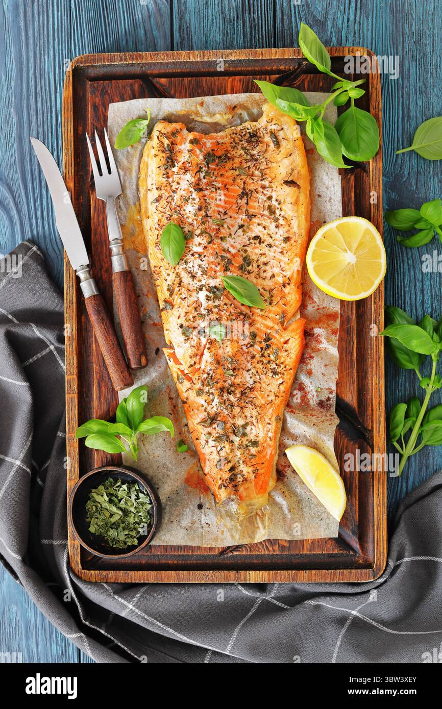 Gegrillte ganze Lachsfilets mit Haut auf einem rustikalen Holzschneidebrett mit frischem Basilikum, Zitronenscheiben, Besteck und Gewürzen auf Holztisch, vertica Stockfoto