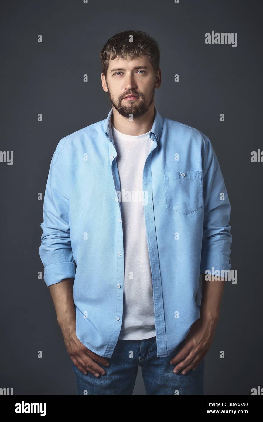 Dieser junge selbstbewusste Mann mit Bart in stilvoller Freizeitkleidung posiert im Studio vor grauem Hintergrund. Porträt eines gutaussehenden Mannes aus Kaukasien Stockfoto