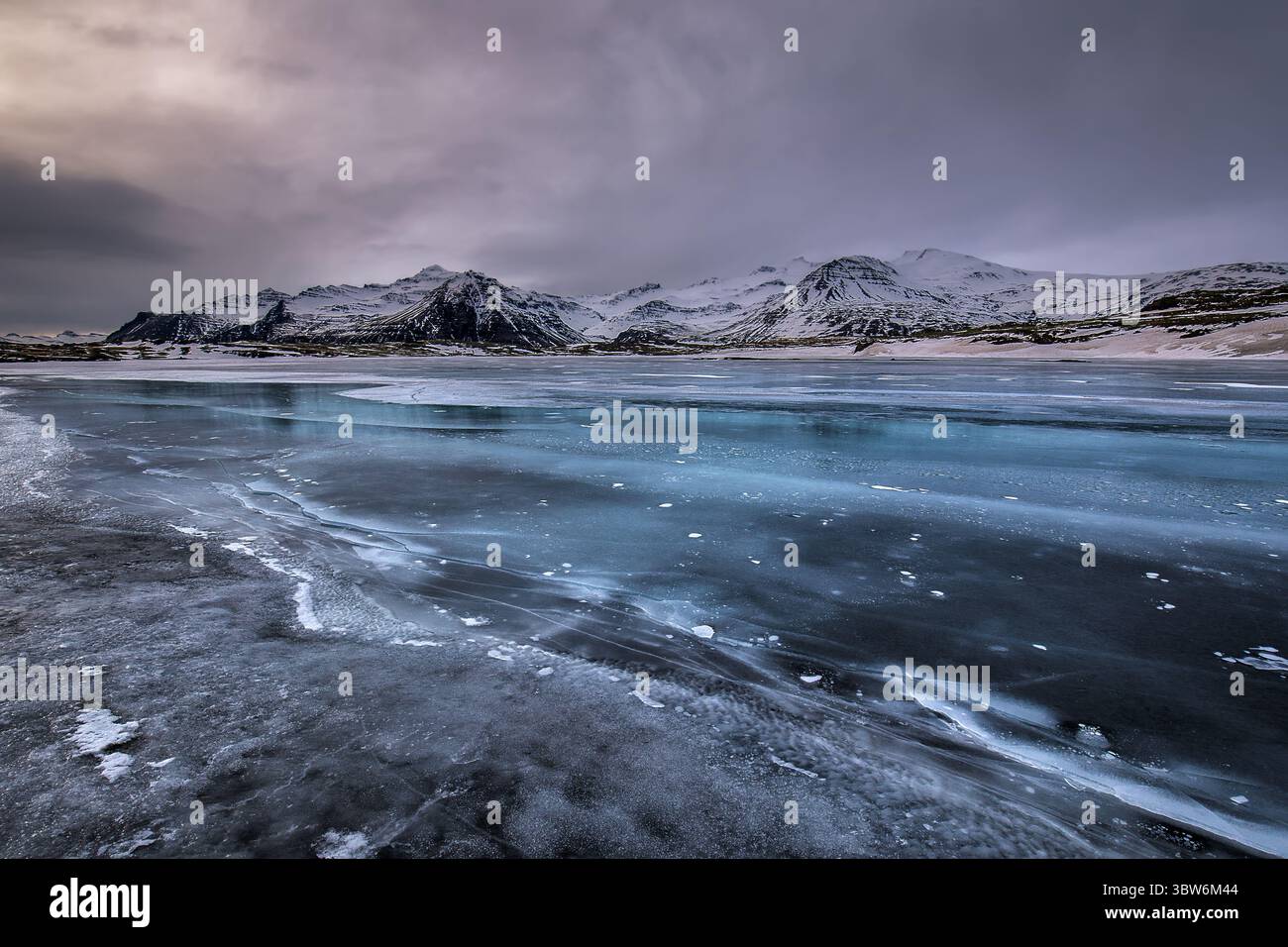 Blick auf eisige Muster tanzen über den teilweise gefrorenen See und reflektieren die schneebedeckten Berge unter einem strukturierten Himmel, Vik, Island. Stockfoto