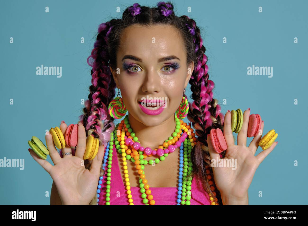 Nahaufnahme einer schillernden Maiden mit einem bunten Zöpfe Frisur und Make-up, im Studio vor einem blauen Hintergrund posiert, holding Maca Stockfoto