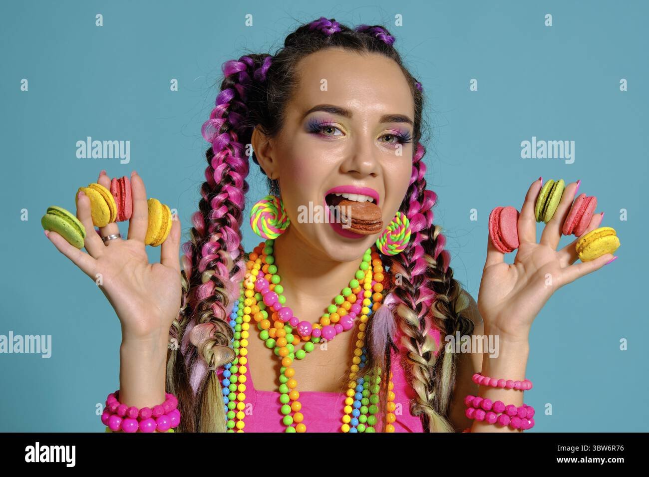 Nahaufnahme einer glücklich Maiden mit einem bunten Zöpfe Frisur und Make-up, im Studio vor einem blauen Hintergrund posiert, holding Macaron Stockfoto