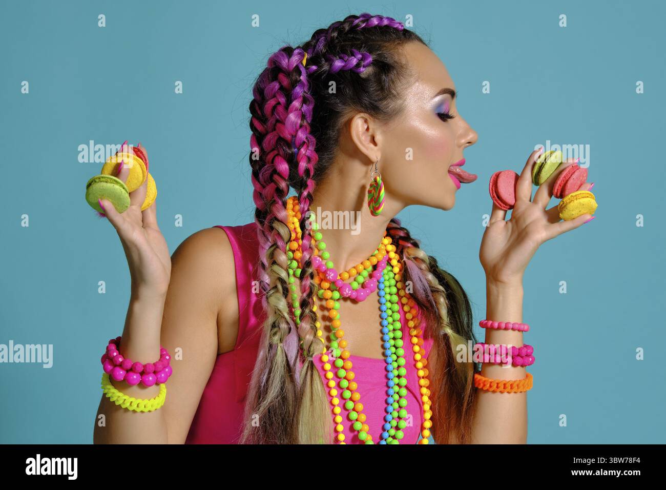 Nahaufnahme einer reizenden Dame mit einem bunten Zöpfe Frisur und Make-up, im Studio vor einem blauen Hintergrund posiert, holding Maca Stockfoto