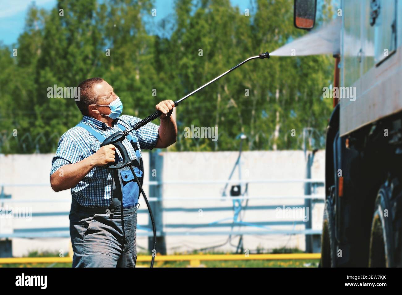 Autowaschanlage für Sonderausrüstung und LKW. Ein männlicher Fahrer mit einer medizinischen Maske wäscht im Sommer unter Druck auf der Straße einen Lastwagen mit einer Pistole Stockfoto