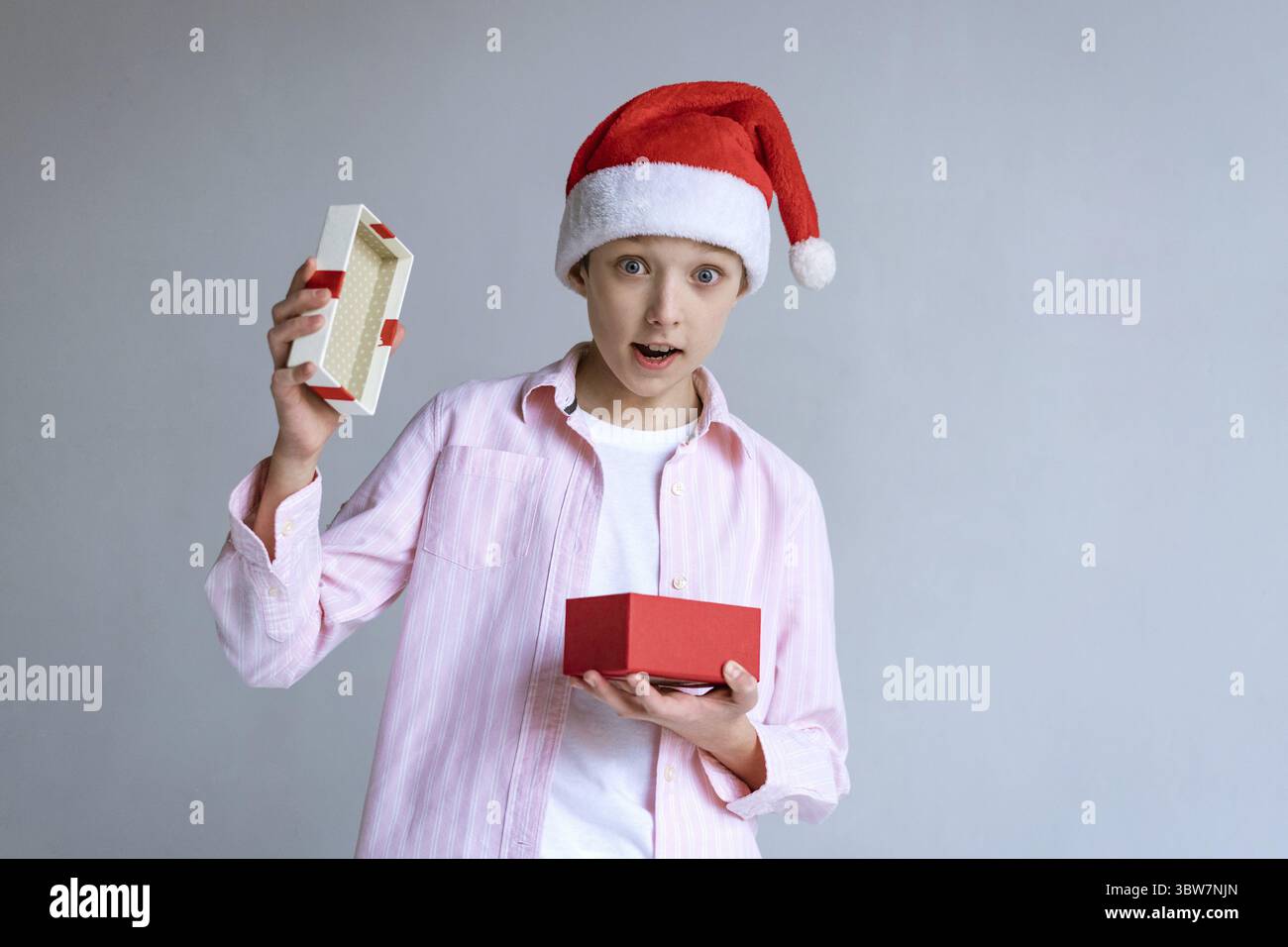 Kaukasier in santa claus Hut und Hemd öffnet Geschenk und sieht überrascht auf einem hellen Hintergrund im Studio aus. Happy Boy erhielt eine Kiste mit einem GIF Stockfoto