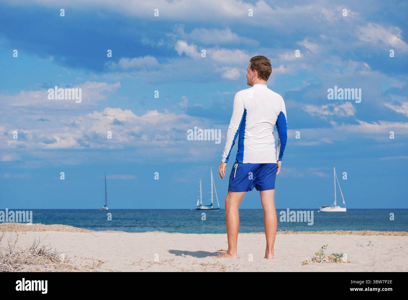 Rückansicht eines Mannes, der in einem sonnengeschützten Hemd am Strand steht und auf das Meer und die Segelboote blickt. Sommerurlaub und Wassersportkonzept. Stockfoto