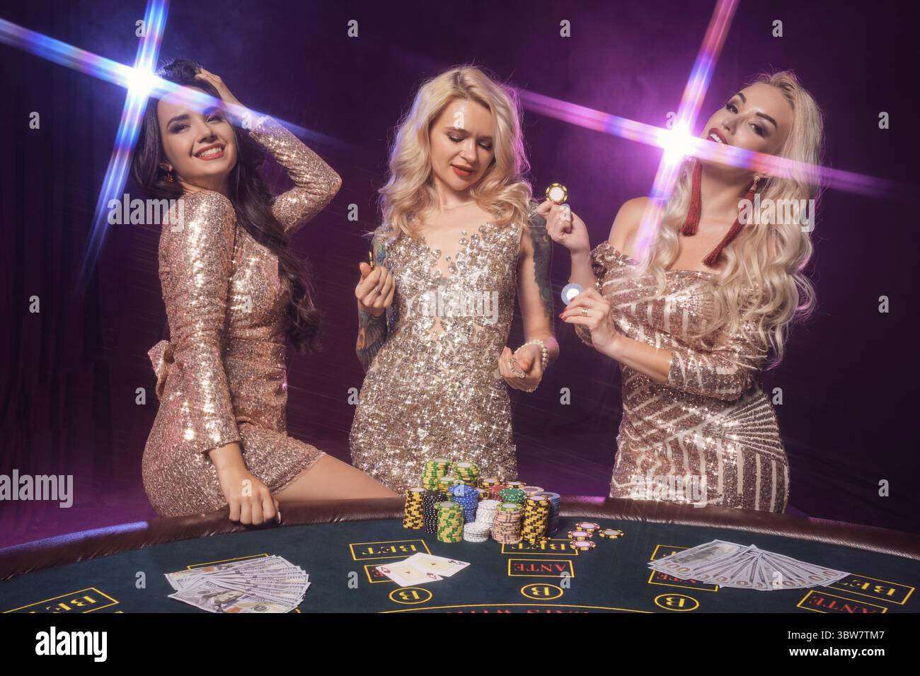 Schöne Frauen mit perfekter Frisur und leuchtendem Make-up, gekleidet in goldenen, glänzenden Kleidern, posieren an einem Spieltisch. Pokerkonzept Stockfoto
