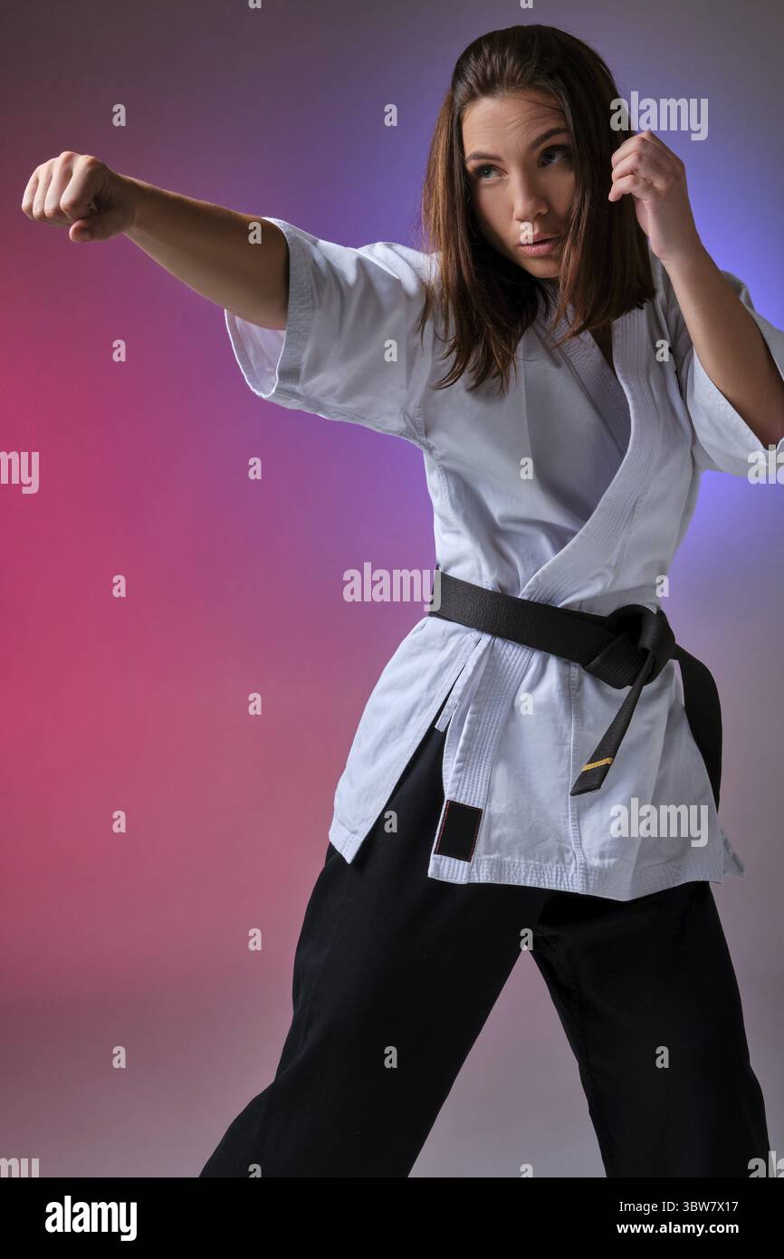 Junge starke Jungfrau mit lockeren Haaren, gekleidet in einem traditionellen Kimono, schaut weg, während sie Karate vor einem farbenfrohen Studio-Hintergrund übt. März Stockfoto