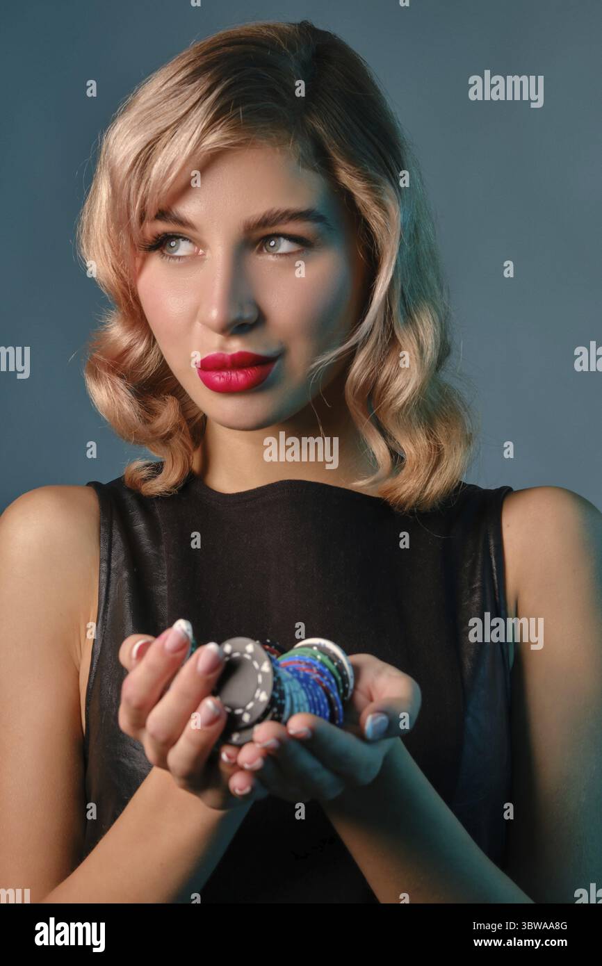 Schöne blonde Jungfrau mit hellem Make-up, in schwarzem Lederkleid hält ein paar bunte Chips und posiert vor blauem Studiohintergrund. Glücksspiel Stockfoto
