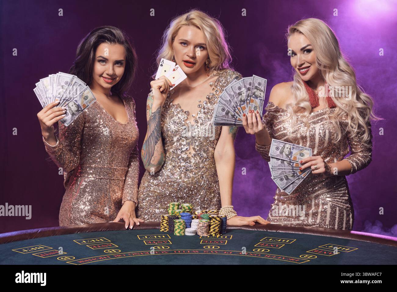 Schöne Damen mit perfekter Frisur und leuchtendem Make-up, gekleidet in goldenen, glänzenden Kleidern, posieren vor einem Spieltisch und sehen aus wie eine Stockfoto