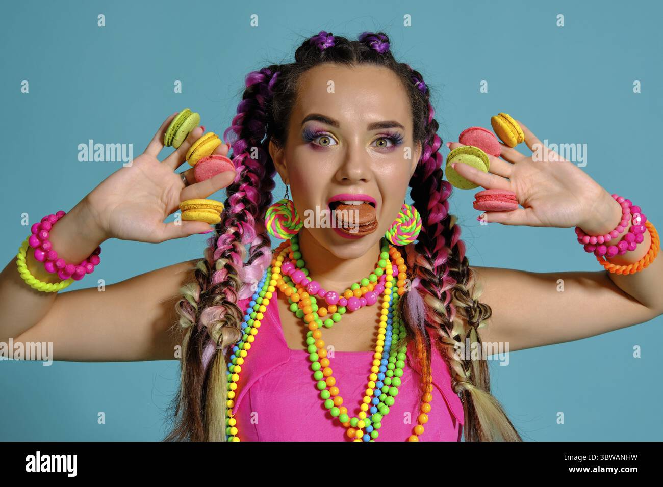 Nahaufnahme einer glücklichen Dame mit einem bunten Zöpfe Frisur und Make-up, im Studio vor einem blauen Hintergrund posiert, holding Macaron i Stockfoto