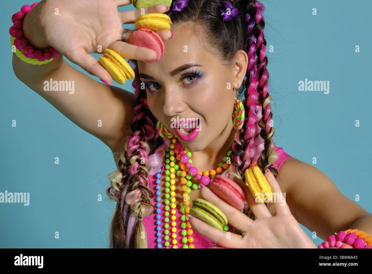 Nahaufnahme einer entzückenden Mädchen mit einem bunten Zöpfe Frisur und Make-up, im Studio vor einem blauen Hintergrund posiert, holding Maca Stockfoto
