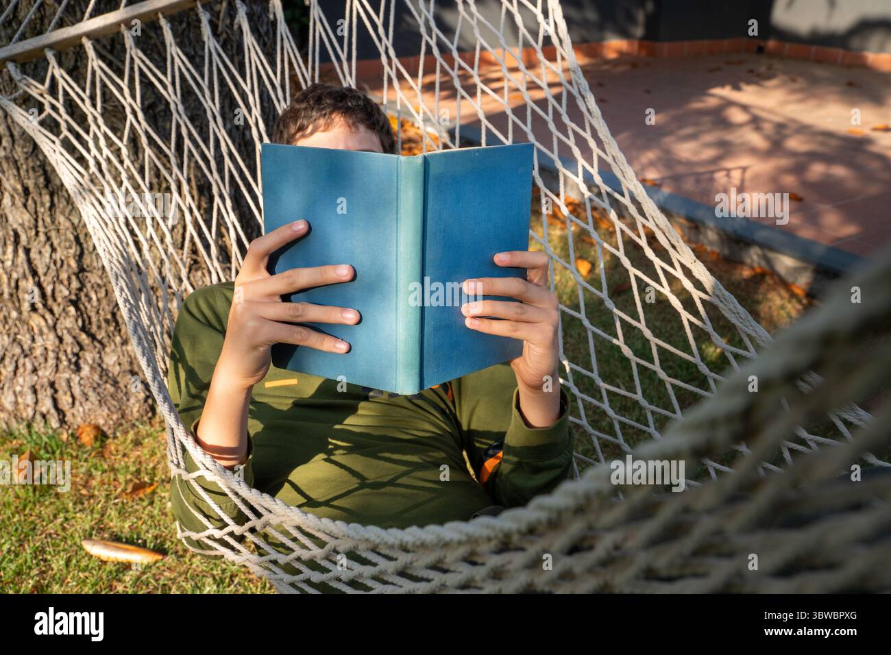 Eine Person versteckt ihr Gesicht hinter einem Buch, während sie sich in einer Hängematte im Freien entspannt, ein kraftvolles Konzept von Eskapismus, Fantasie und der Privatsphäre des Lesens Stockfoto