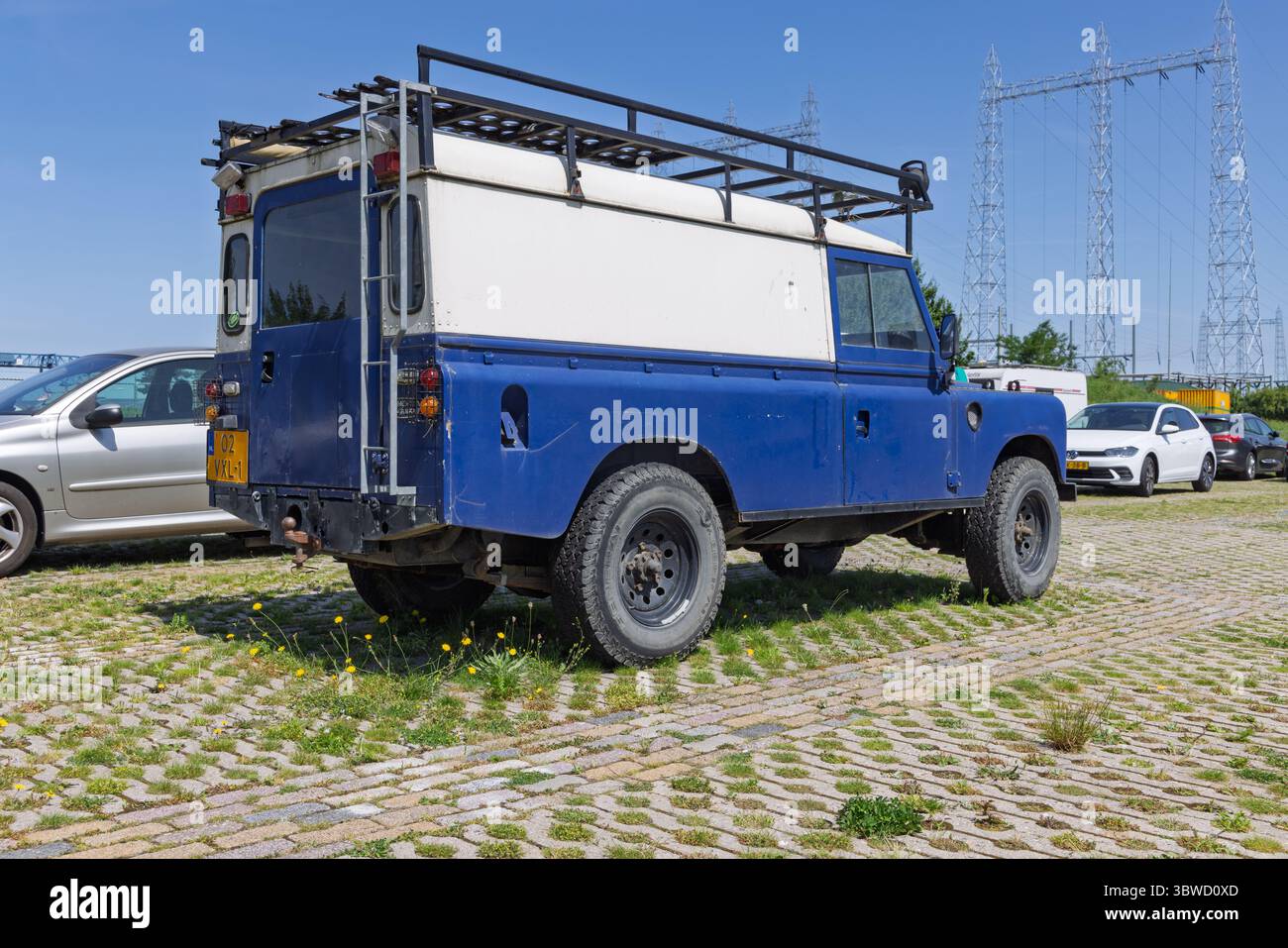 Rückansicht eines klassischen blauen Land Rover der Serie 3 mit Dachträger, der auf einem gepflasterten Gelände geparkt ist und im Hintergrund Stromleitungen aufweist. Nijmegen, Niederlande, 2 Stockfoto