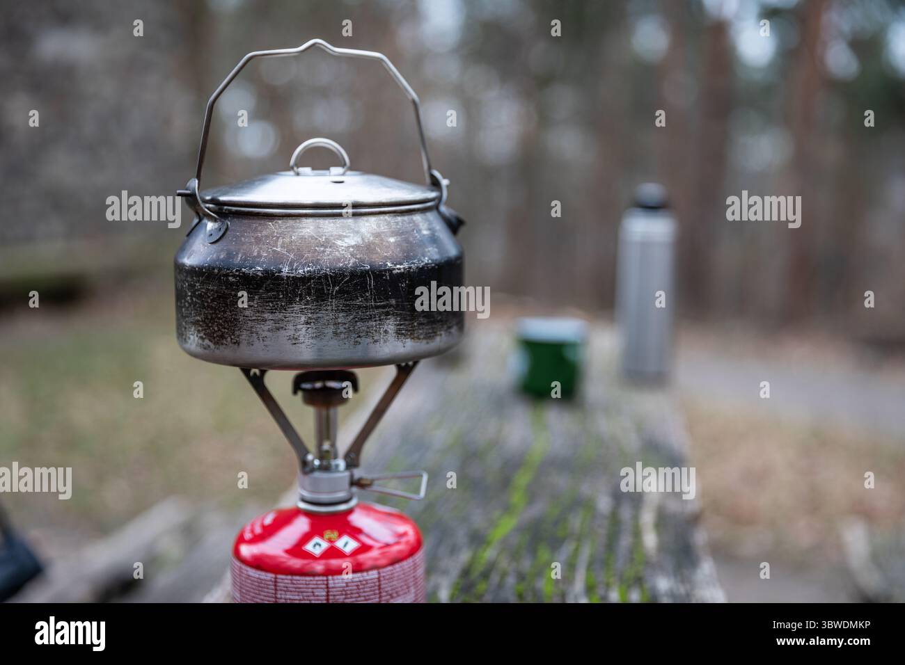 Abgenutzter Metallkessel auf Gasherd auf verwittertem Holztisch im Freien, verschwommener Campingbecher, Thermos und Waldweg im Hintergrund, Fokus auf den Wasserkocher. Stockfoto