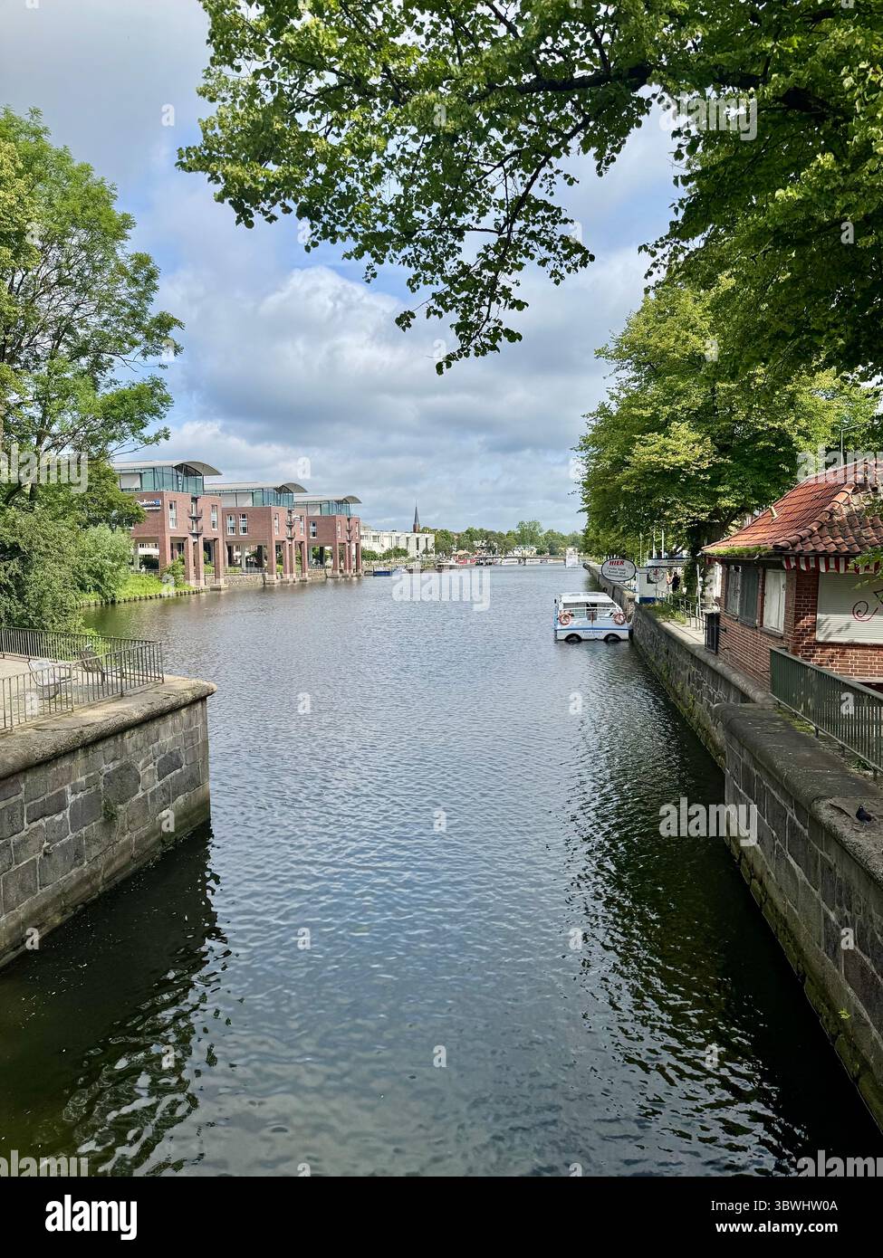 Friedlicher Blick auf den Kanal in lübeck, gesäumt von historischer und moderner Architektur, grünen Bäumen und einem Boot am Anleger unter einem bewölkten Sommerhimmel. - Smartphone-aufgenommenes Stockfoto
