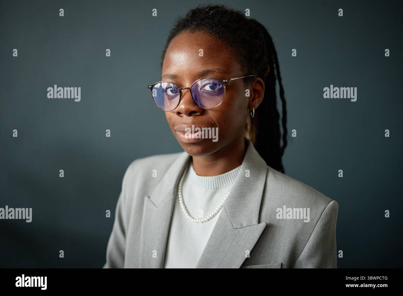 Porträt einer jungen erwachsenen Schwarzen Frau, die Brillen und Schmuck trägt und mit selbstbewusstem Ausdruck in die Kamera blickt, in professioneller Kleidung vor einem einfachen Hintergrund steht Stockfoto