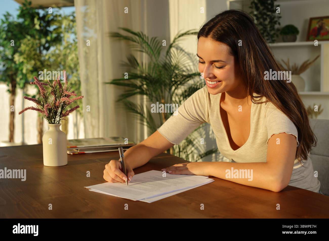 Glückliche Frau, die einen Vertrag auf Holztisch bei Sonnenuntergang zu Hause unterschreibt Stockfoto
