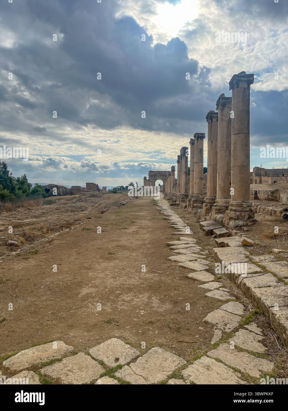 Alte römische Kolonnade und Bogengang in Jerash, Jordanien, unter dramatischem bewölktem Himmel, mit unbefestigtem Pfad und Überresten von Steinpflaster. Stockfoto