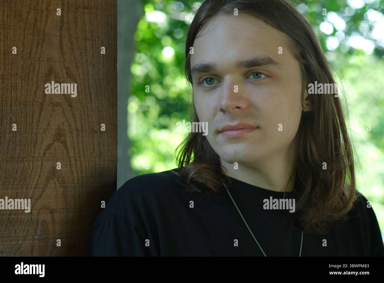 Ein junger Mann steht nachdenklich an einer hölzernen Säule, umgeben von Grün. Stockfoto