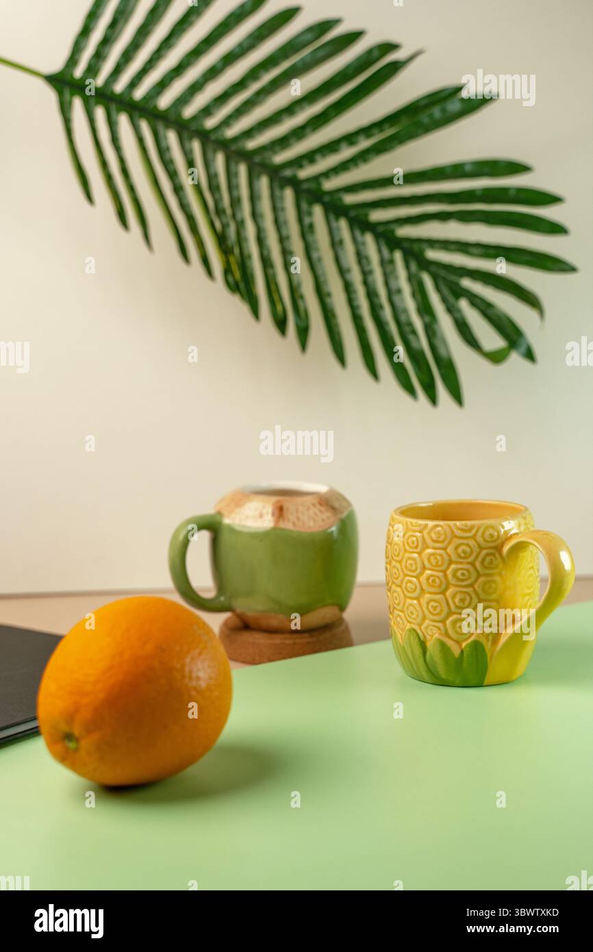 Zwei lustige, tropische Tassen in Form einer grünen Kokosnuss und einer gelben Ananas. Frische Orange und ein Palmblatt sorgen für einen Sommerurlaub. Stockfoto