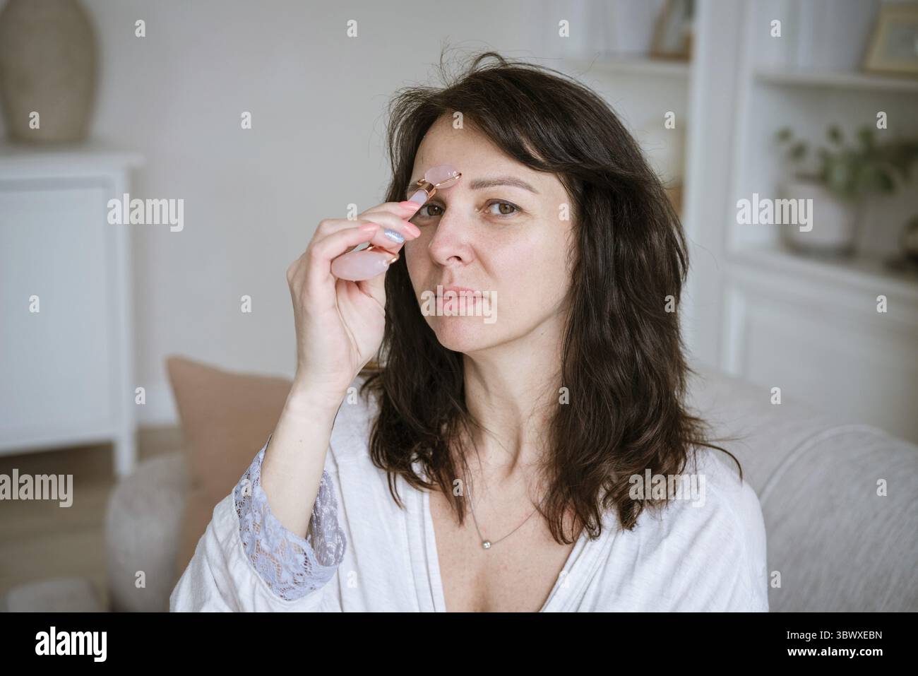 Porträt einer kaukasischen Frau, die Gesichtsmassage mit Quarzschaber für rosa Gouache macht, trägt einen Bademantel, der auf Sofa im gemütlichen Wohnzimmer zu Hause sitzt, Anti Stockfoto