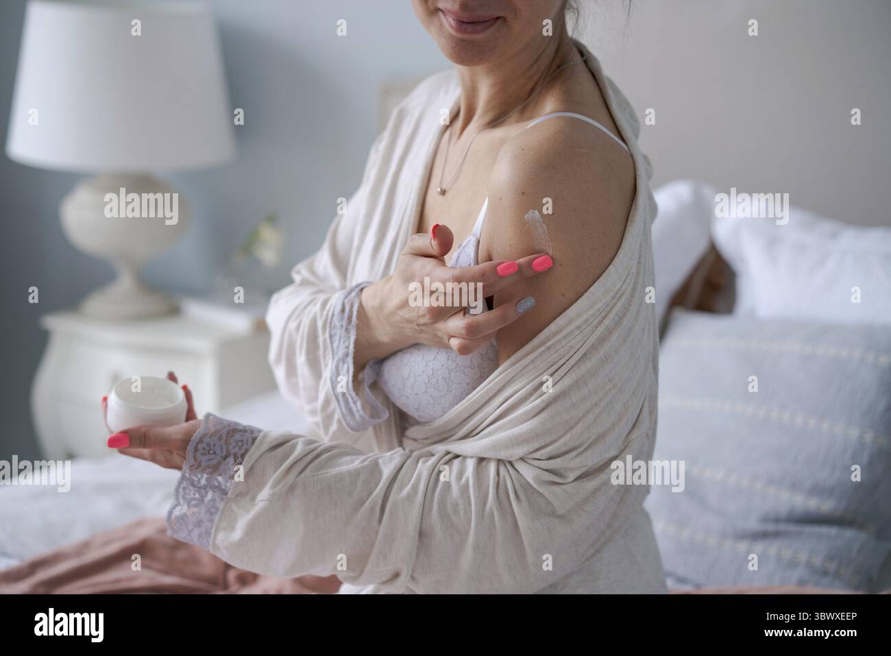 Junge Frau im Hausmantel, die natürliche handgemachte Kosmetikprodukte auf ihre Hände aufträgt, während sie morgens auf dem Bett sitzt. Hautpflege zu Hause durch kaukasier g Stockfoto