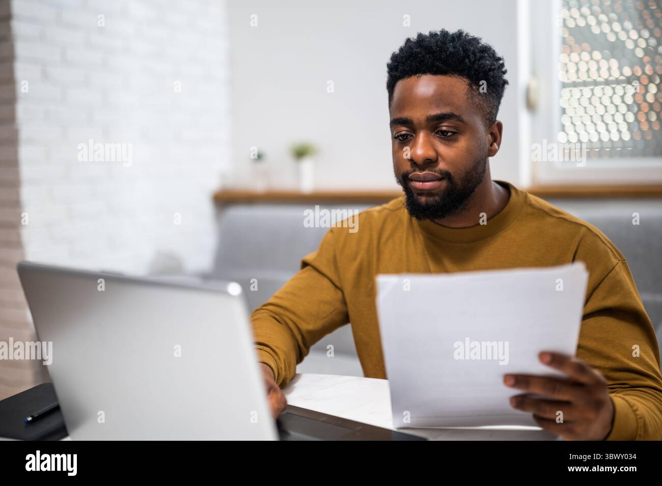 Junger afrikanischer Geschäftsmann, der auf Laptop schreibt und Dokumente im Wohnzimmer analysiert. Er arbeitet von zu Hause aus. Stockfoto
