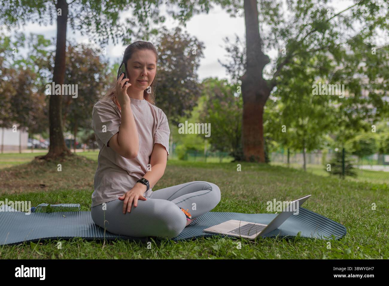 Frau sitzend im Kreuz im Freien sprechendes Smartphone hält Gerät zu Ohr blasses Hemd graue Leggings offenes Notebook Silber Laptop grüner Gras Hintergrund Stockfoto