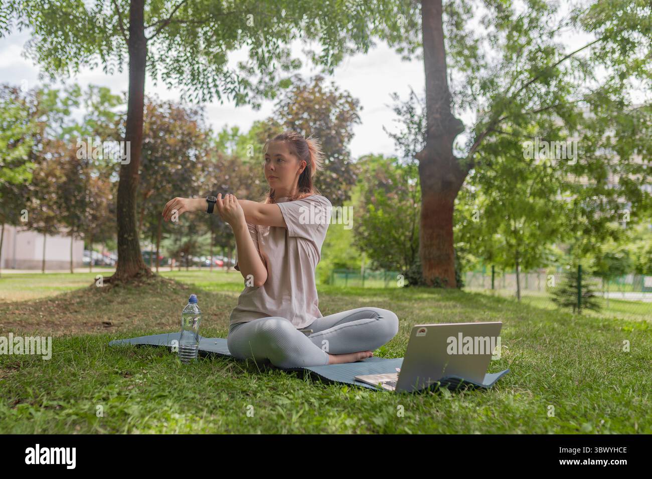 Frau dehnt ihren Arm, während sie auf einer Yogamatte mit Laptop sitzt, während sie im Freien trainieren. Stockfoto