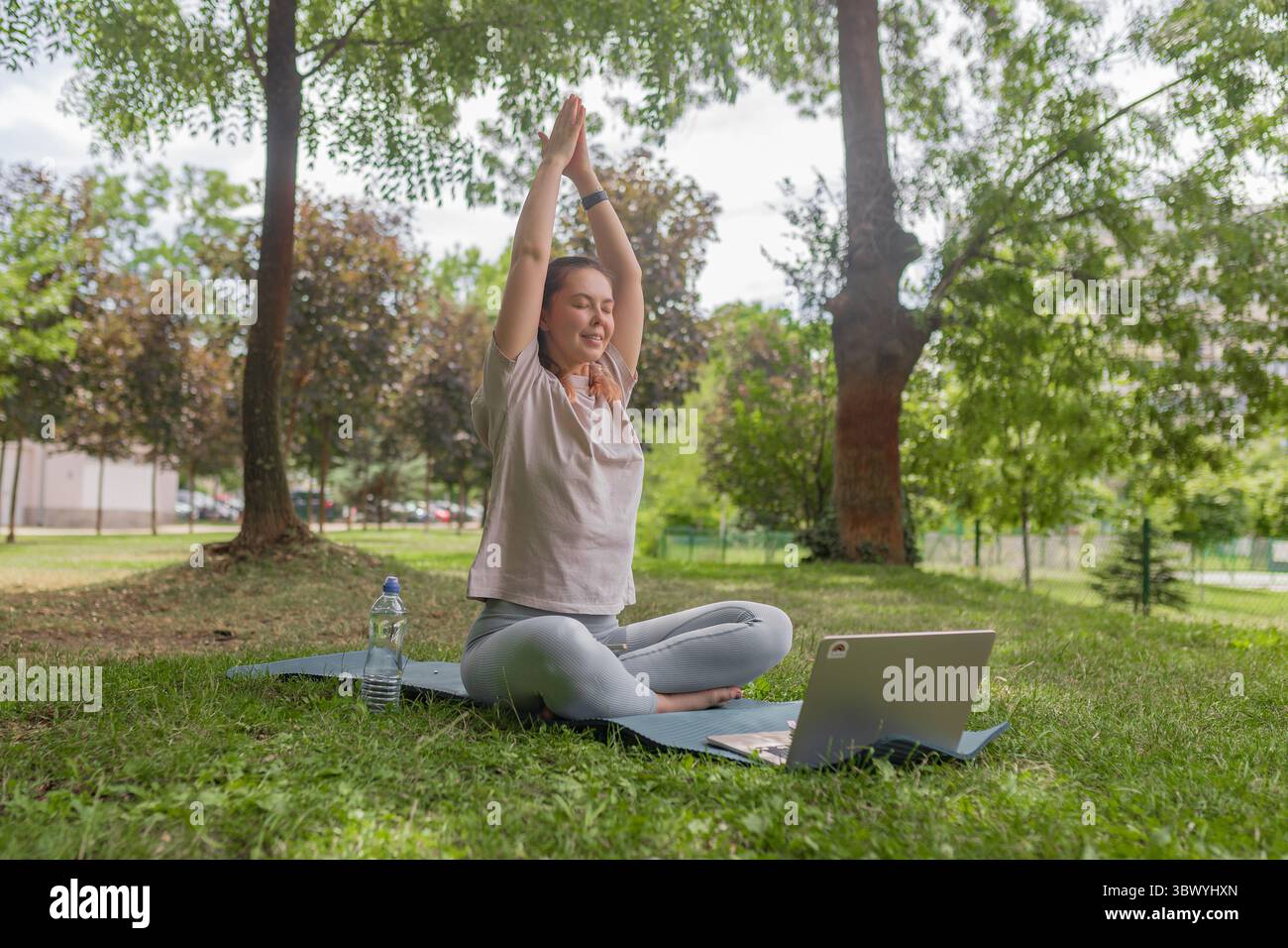 Frau, die die Arme in aufsteigender Yoga-Pose während der Übung im Freien mit Laptop im Park hebt. Stockfoto