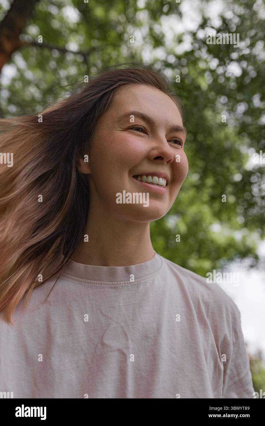 Junge Frau, die draußen lächelt und ein hellrosa T-Shirt trägt, umgeben von grünem grünem Hintergrund. Stockfoto