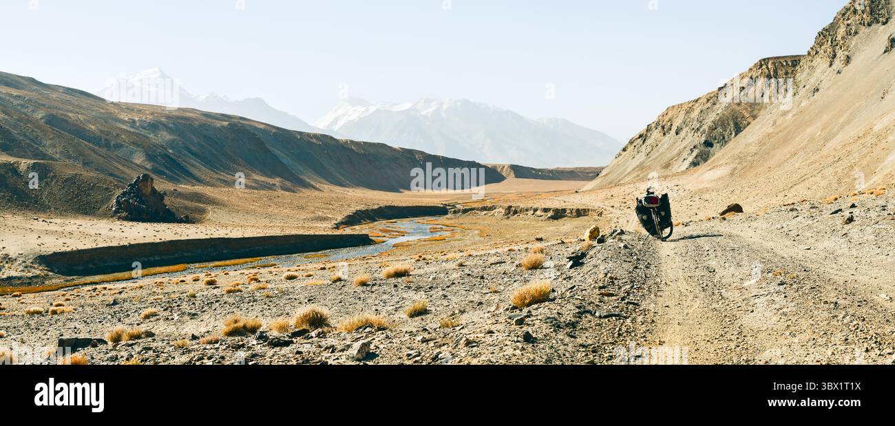 Rotes Tourenrad mit schwerem Gepäck in vier Gepäcktaschen steht an der Schotterstraße, umgeben von wilder, ariden Pamir-Bergen, alpiner Natur Stockfoto