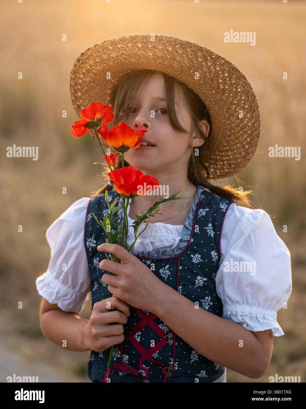 Nahaufnahme eines entzückenden Mädchens in Dirndl und Strohhut mit rotem Mohn in der Nähe ihres Gesichts. Traditionelle Volkstracht, Kinderunschuld, natürlicher Sommer bea Stockfoto