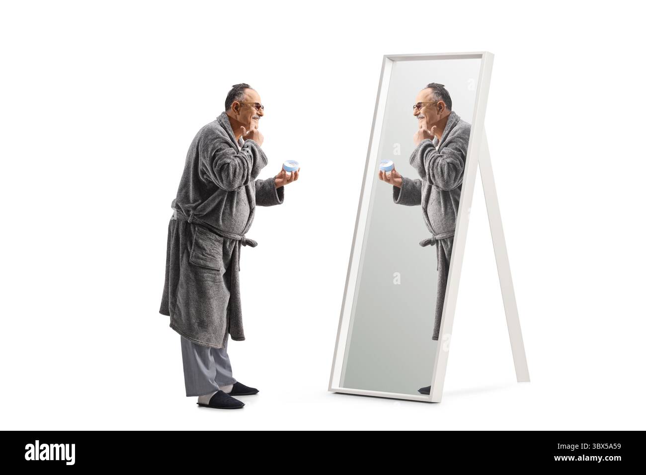 Reifer Mann in einem Bademantel, der Gesichtscreme vor einem Spiegel auf weißem Hintergrund aufträgt Stockfoto
