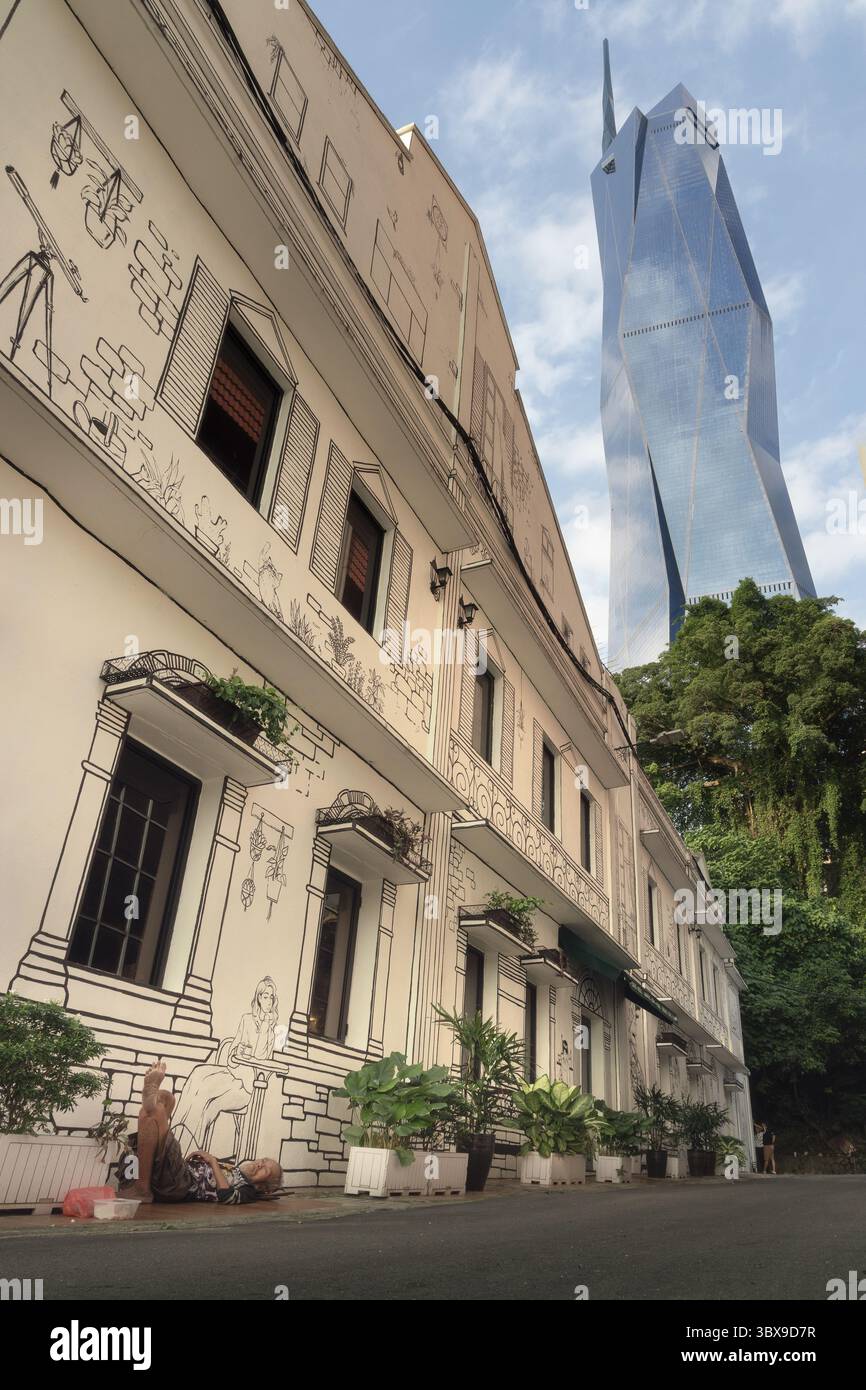 In Kuala Lumpurs Chinatown liegt ein Obdachloser unter einem kulturell bemalten Gebäude, hinter dem sich die hoch aufragende Merdeka 118 erhebt. Stockfoto