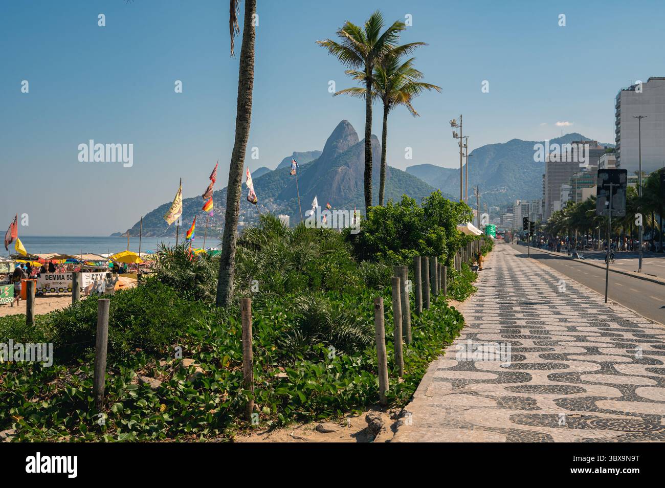 Ikonischer Mosaik-Gehweg der Copacabana in Rio de Janeiro mit Palmen, farbenfrohen Strandflaggen und einem malerischen Blick auf Ipanema, Leme Hill und Sugarloaf MoU Stockfoto