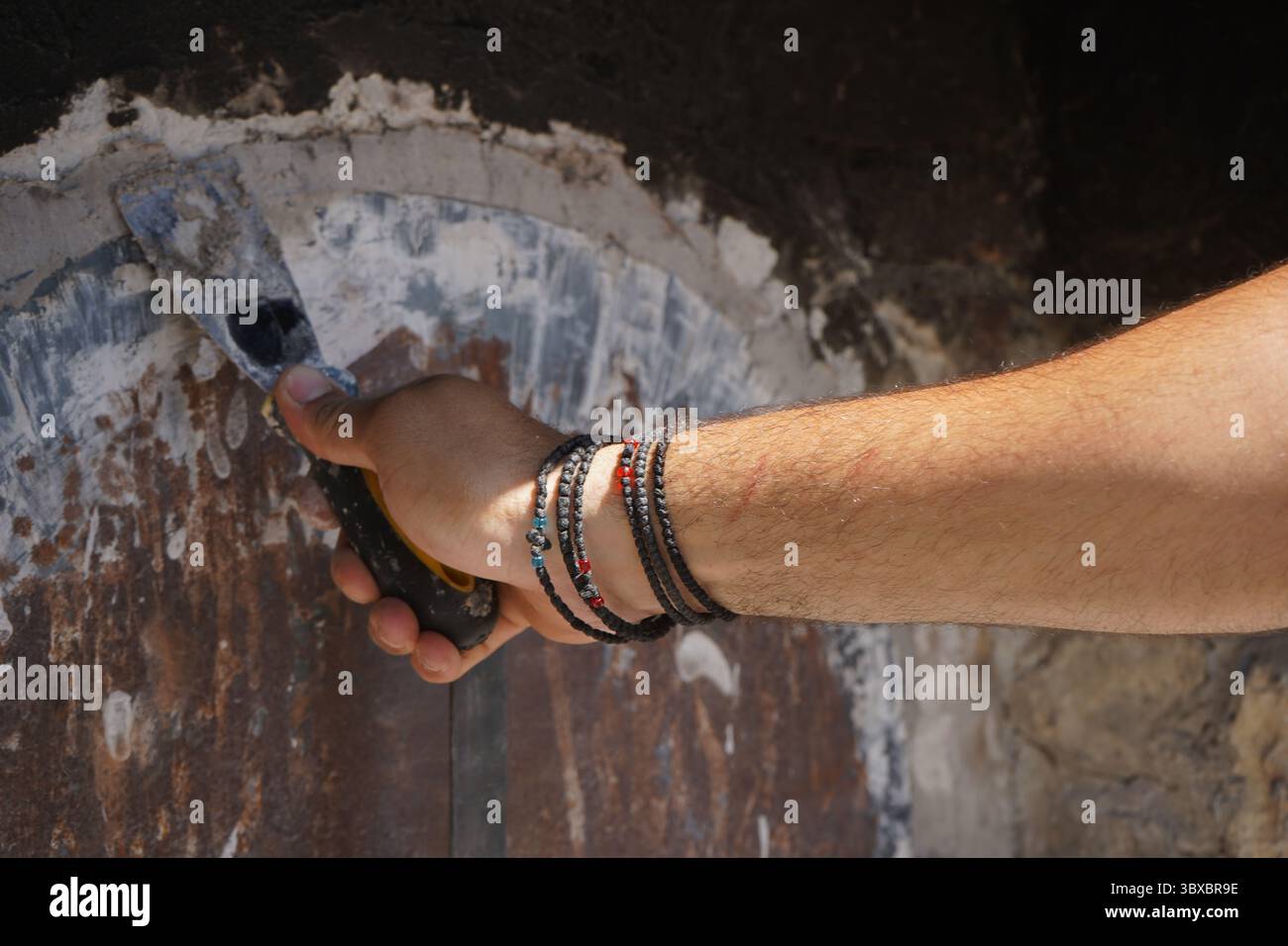 Die Hand eines jungen Mannes versiegelt eine holzbefeuerte Ofentür mit einem Spachtel und einer Aschemischung. Im Inneren warten Trays mit traditionellen kretischen Gerichten auf langsames Backen. Stockfoto