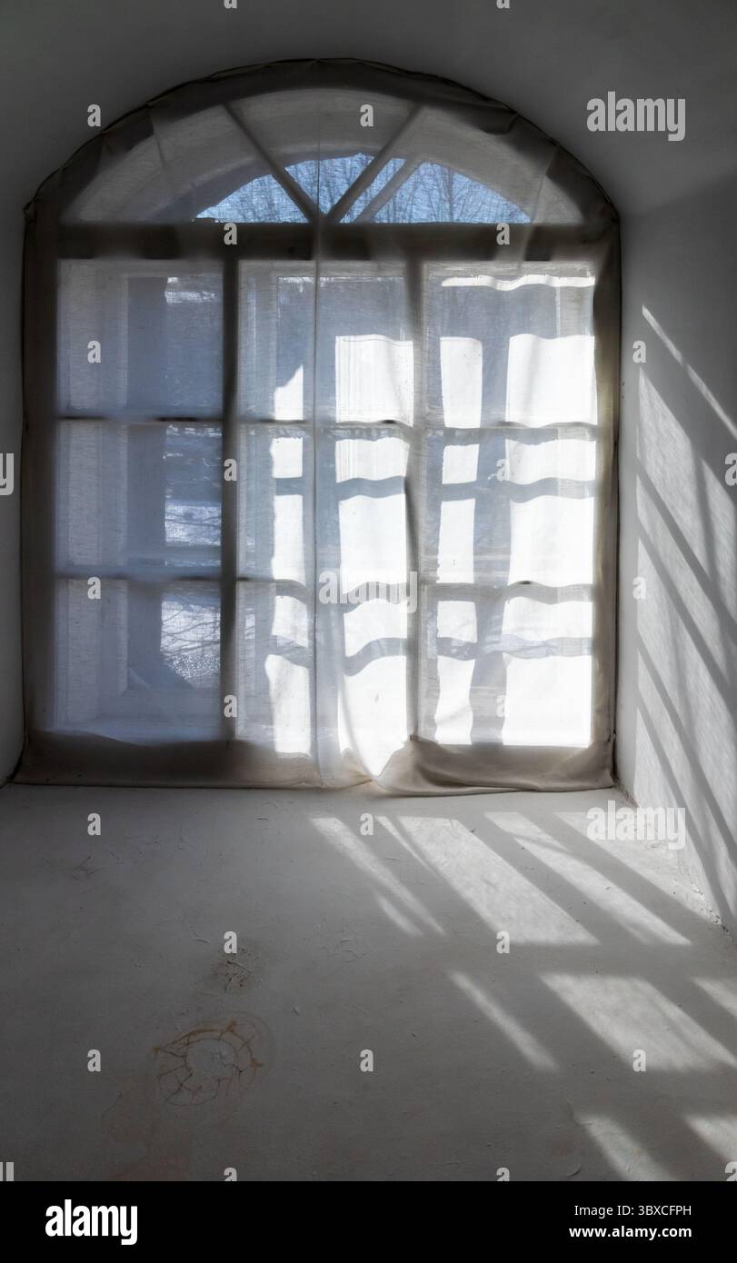 Ein sanft beleuchtetes Fenster mit transparenten Vorhängen und Sonnenstrahlen, die Schatten werfen. Die ruhige Atmosphäre unterstreicht das Zusammenspiel von Licht und Texturen und schafft Stockfoto