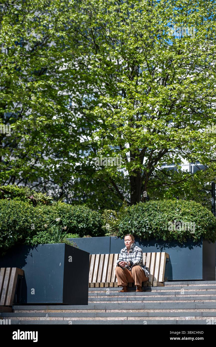 An einem hellen Frühlingstag sitzt eine Person allein auf einer Bank, umgeben von Grün und Bäumen in Canary Wharf, London. Stockfoto