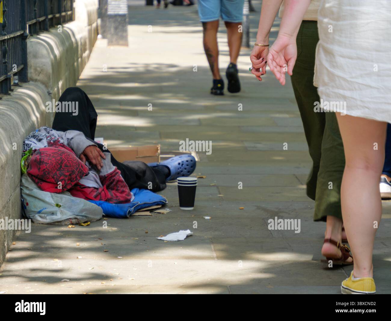 Fußgänger gehen an einem Obdachlosen vorbei, der auf einer belebten Londoner Straße neben einem Becher für Spenden liegt, was die zunehmende Sichtbarkeit von Obdachlosigkeit und wirtschaftlicher Not in der britischen Hauptstadt unterstreicht. Stockfoto Fußgänger gehen an einem Obdachlosen vorbei, der auf einer belebten Londoner Straße neben einem Becher für Spenden liegt, was die zunehmende Sichtbarkeit von Obdachlosigkeit und wirtschaftlicher Not in der britischen Hauptstadt unterstreicht. Stockfoto