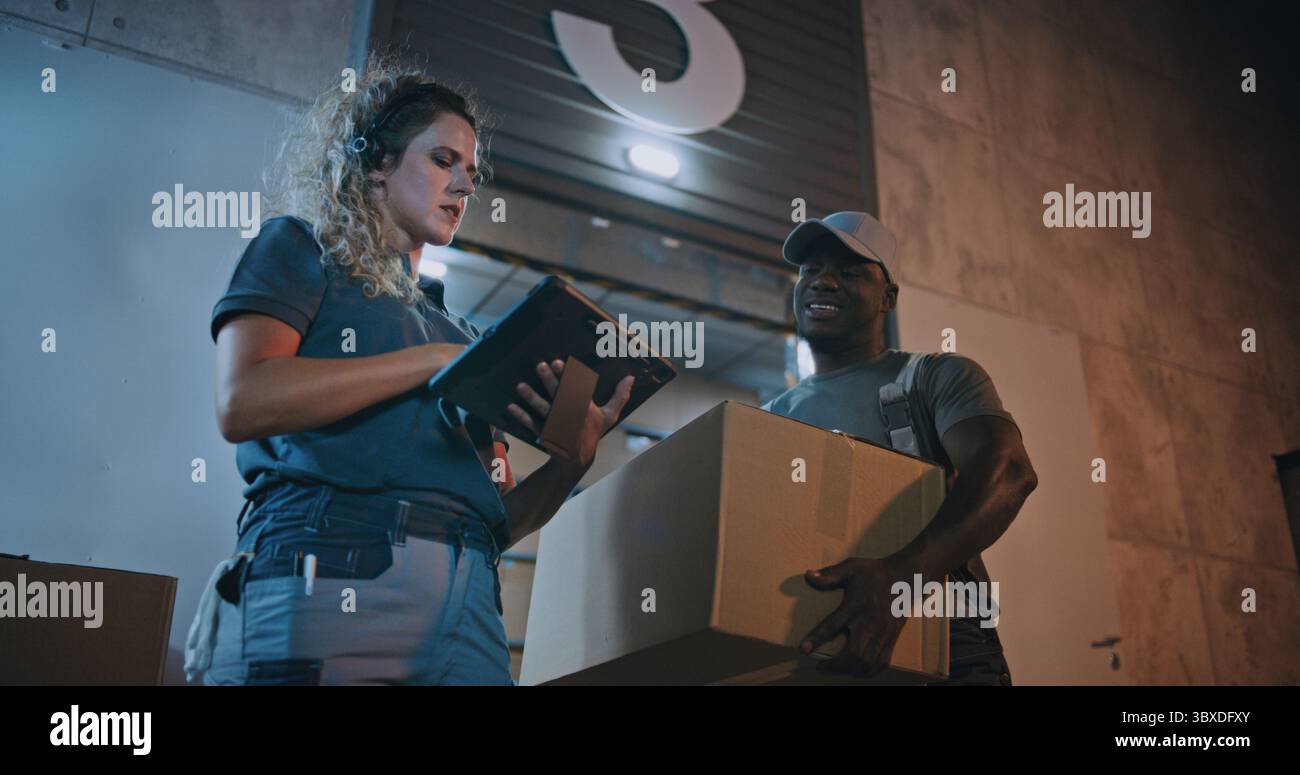 Verschiedene Mitarbeiter, die Kartonschachteln aus dem Logistics Warehouse transportieren und nachts arbeiten. Weibliche Manager scannt Pakete mit Barcode-Scanner mit Digital Tablet. Online-Bestellungen, Lieferung. Zeitlupe. Stockfoto