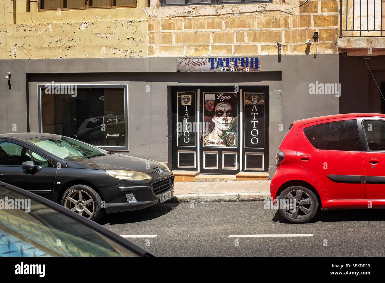Bormla, Malta – 17.07.2025: Steinfassade mit strukturierten Wänden, Tattoostube und geparkten Autos unter der Sonne. Die Atmosphäre des Straßenlebens Stockfoto