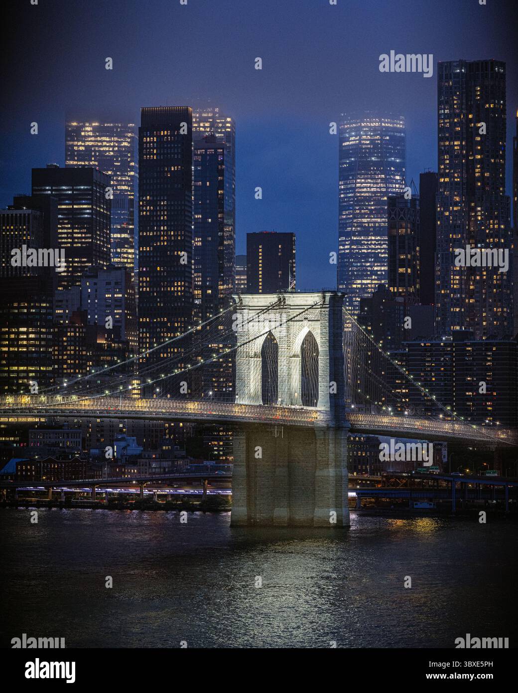 Brooklyn Bridge mit Skyline von New York City bei Nacht – der East River fließt abends mit den Lichtern der Lower Manhattan Wolkenkratzer, die sich in der Abenddämmerung spiegeln Stockfoto