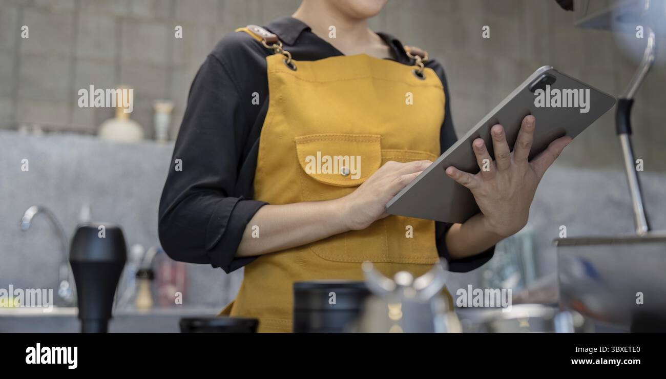 Der Barista trägt eine gelbe Schürze und verwendet ein digitales Tablet in einem modernen Café Stockfoto