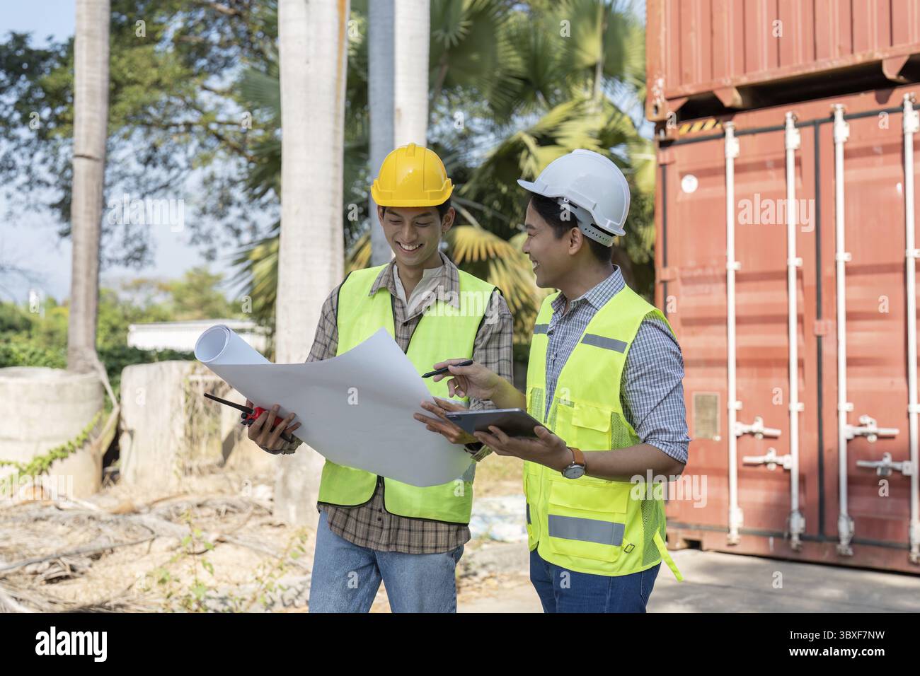 Zwei Ingenieure für Schutzhelme und -Westen lächeln, während sie Logistikpläne überprüfen und an einem Versandcontainer zusammenarbeiten, um Teammitglieder zu präsentieren Stockfoto